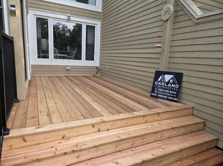 New wood deck with three wide steps beside a house with sage-green siding, with a Garland Contracting sign leaning on the wall