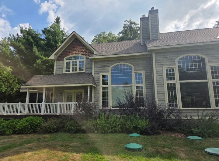 Large cottage-style home with grey wood siding, arched windows, a covered porch with white railings and two chimneys