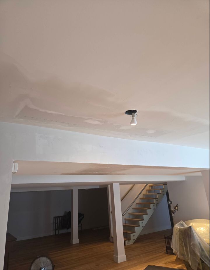 Ceiling with fresh drywall patches above a boxed beam, with an open staircase and hardwood floor below