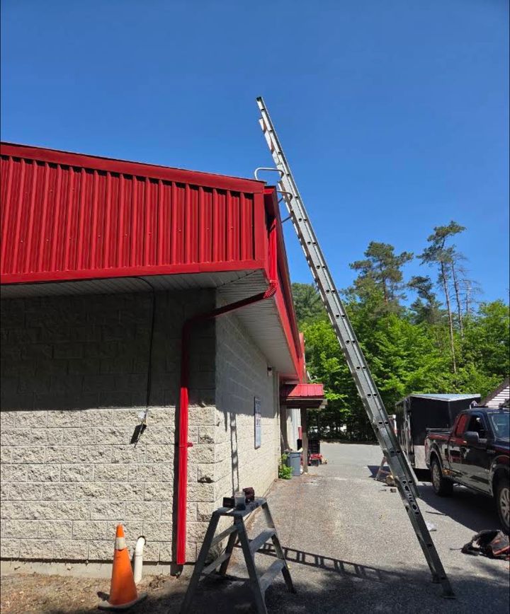 Extension ladder leaning against the red steel roof edge of a block building, with a work truck and trailer parked beside it