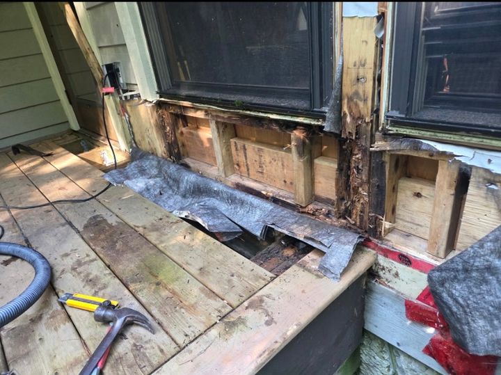 Rotted sill and wall framing exposed under two windows at a deck edge, with old deck boards and tools in the foreground